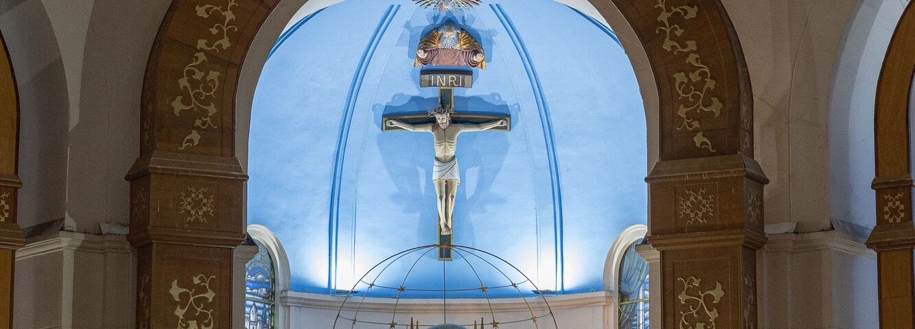 Interior de una iglesia con un crucifijo, vidriera y adornos florales.