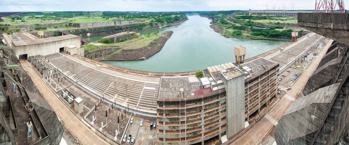 A large dam with a river flowing through it, surrounded by greenery and a parking area.