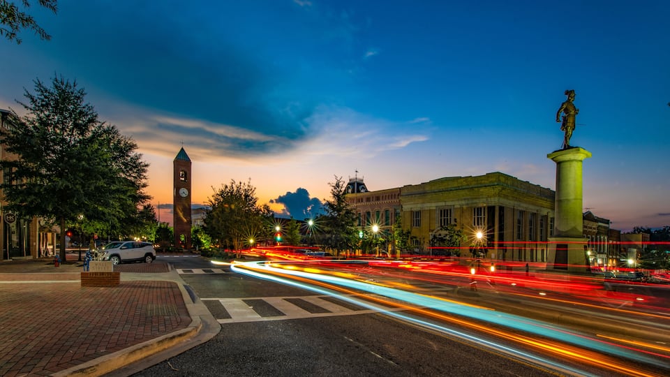 A city street at dusk with a clock tower, a statue, and a building with a dome.