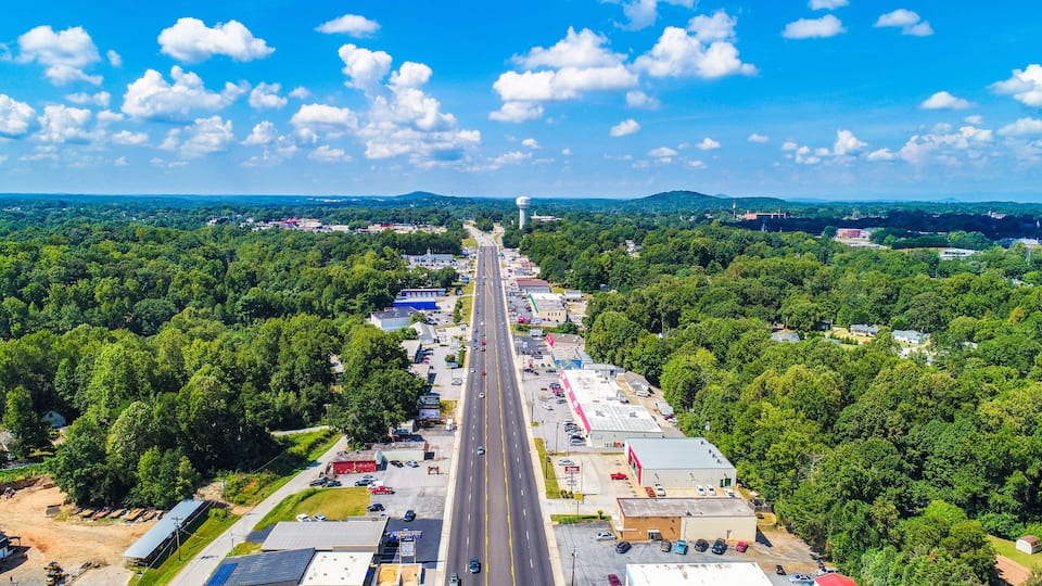 A highway with a clear sky, surrounded by dense green forests and various buildings.