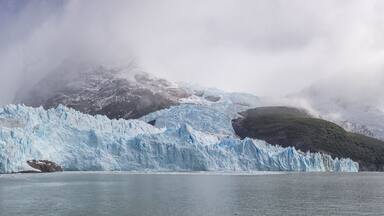 Un glaciar con un gran bloque de hielo desprendiéndose en un fiordo, rodeado de montañas y nubes.