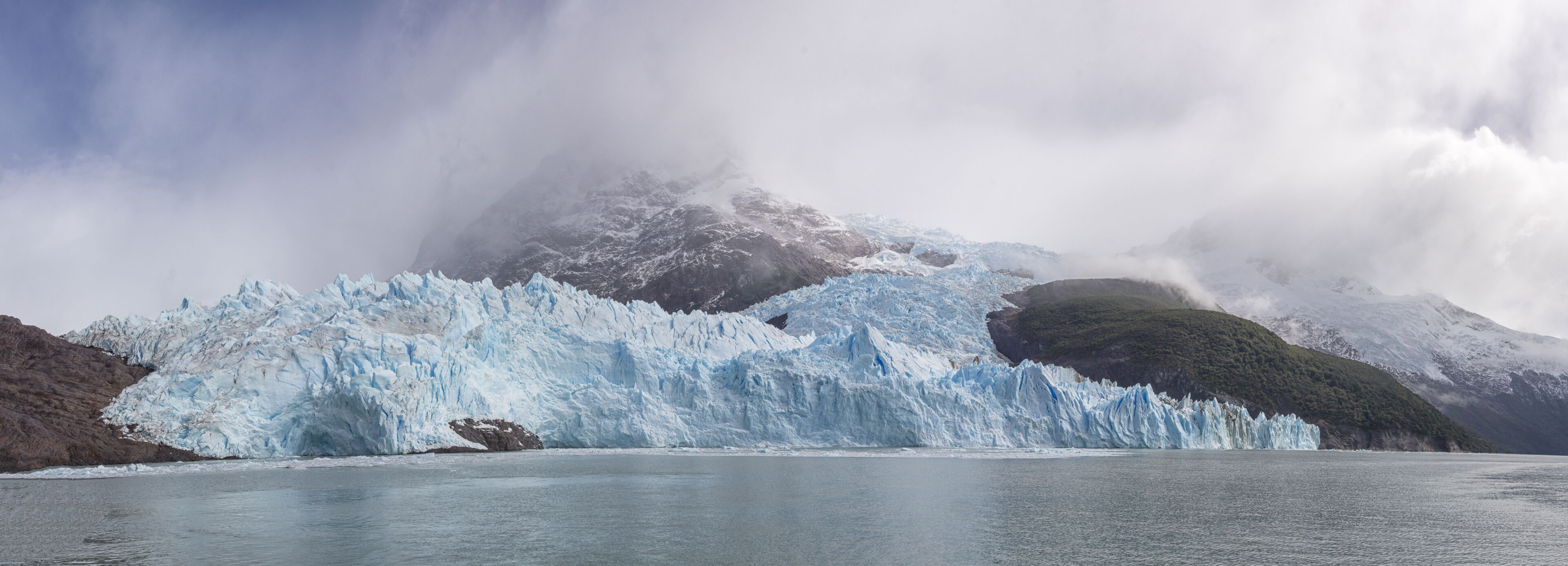 A glacier with a large ice calve in a fjord, surrounded by mountains and clouds.