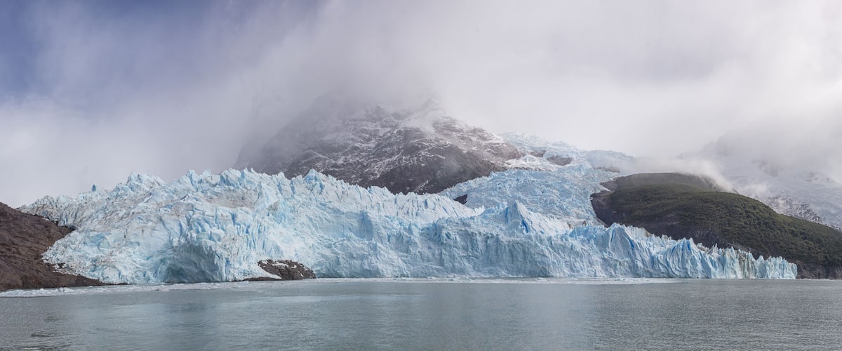 A glacier with a large ice calve in a fjord, surrounded by mountains and clouds.