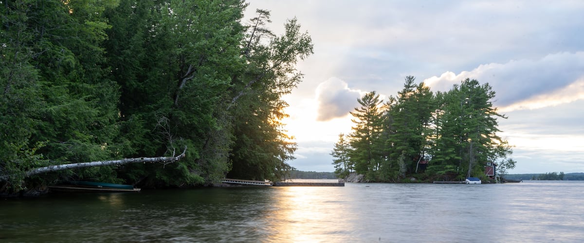 A calm lake with a small island, surrounded by dense trees and a sunset in the background.