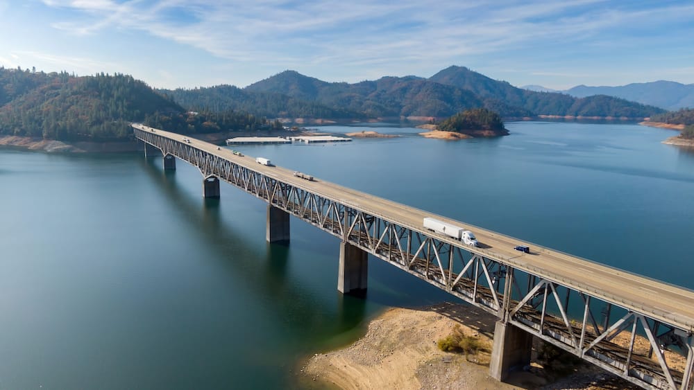 Elevated view of a steel bridge spanning Shasta Lake, with vehicles traversing the roadway. A scenic landscape of mountains and water surrounds the structure. Lakehead, California, USA