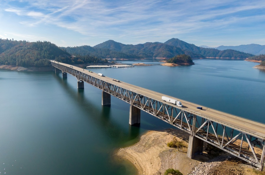 Elevated view of a steel bridge spanning Shasta Lake, with vehicles traversing the roadway. A scenic landscape of mountains and water surrounds the structure. Lakehead, California, USA