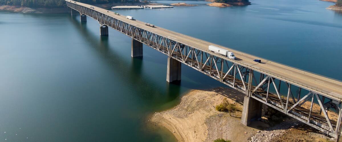A steel bridge over a calm river with mountains in the background.