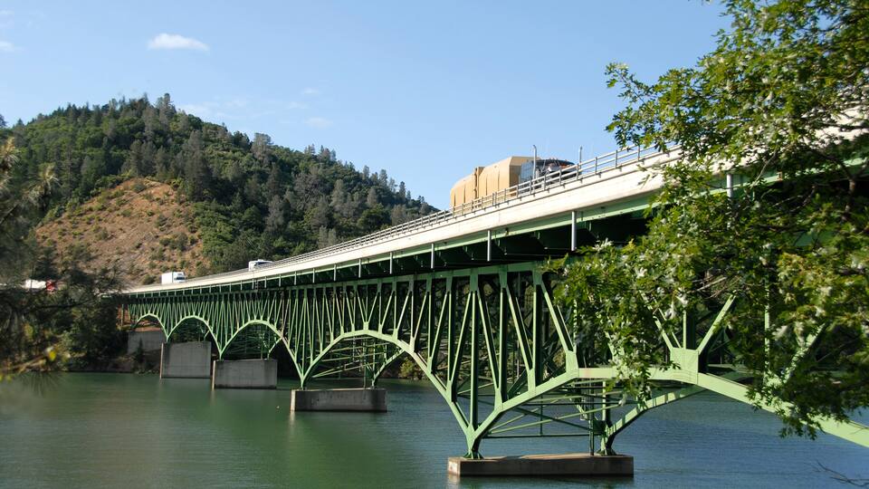 A green metal bridge over a calm river with a hilly landscape in the background.