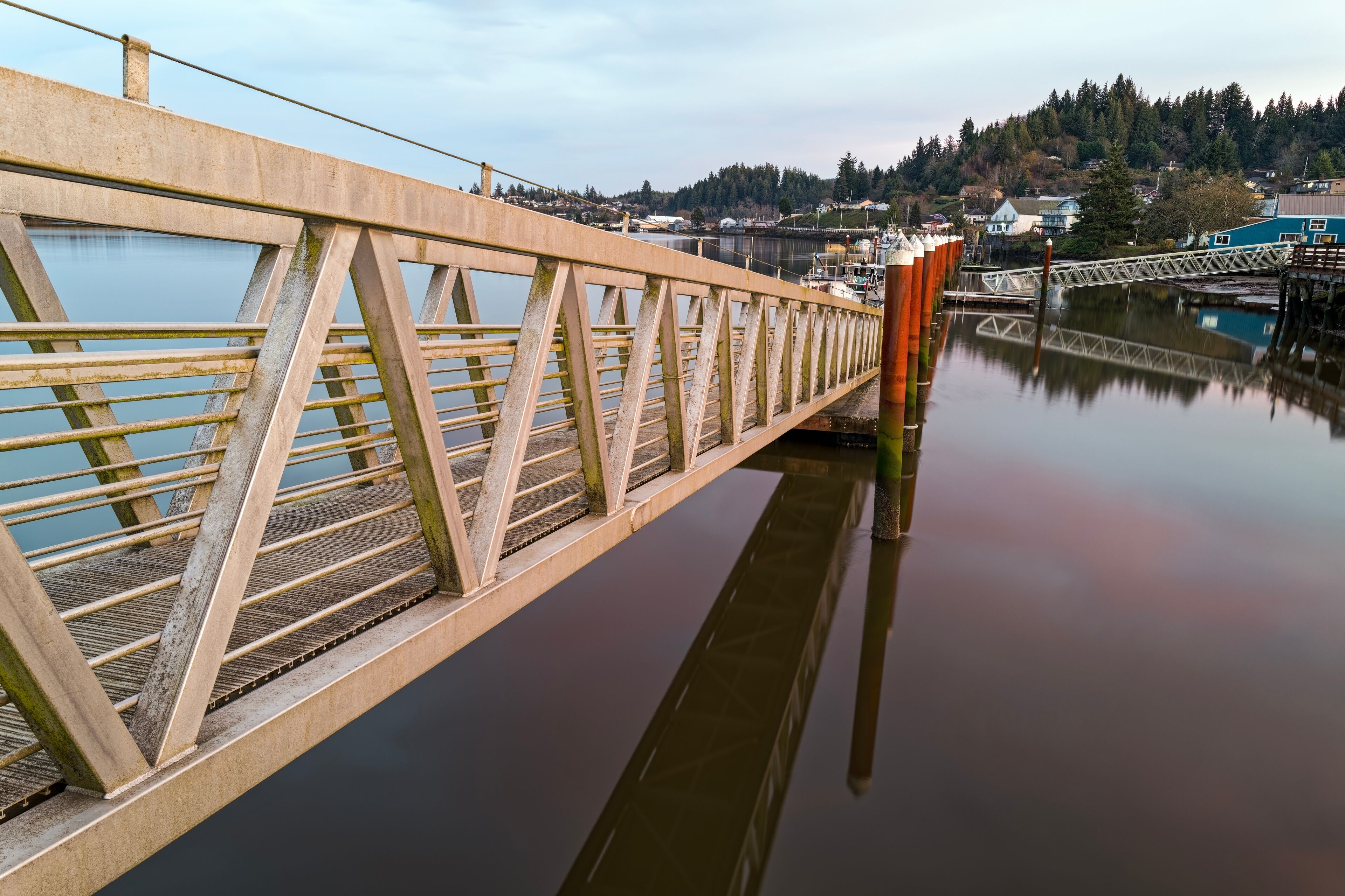 A concrete bridge over calm water with a reflection, a dock, and a forested hill in the background.