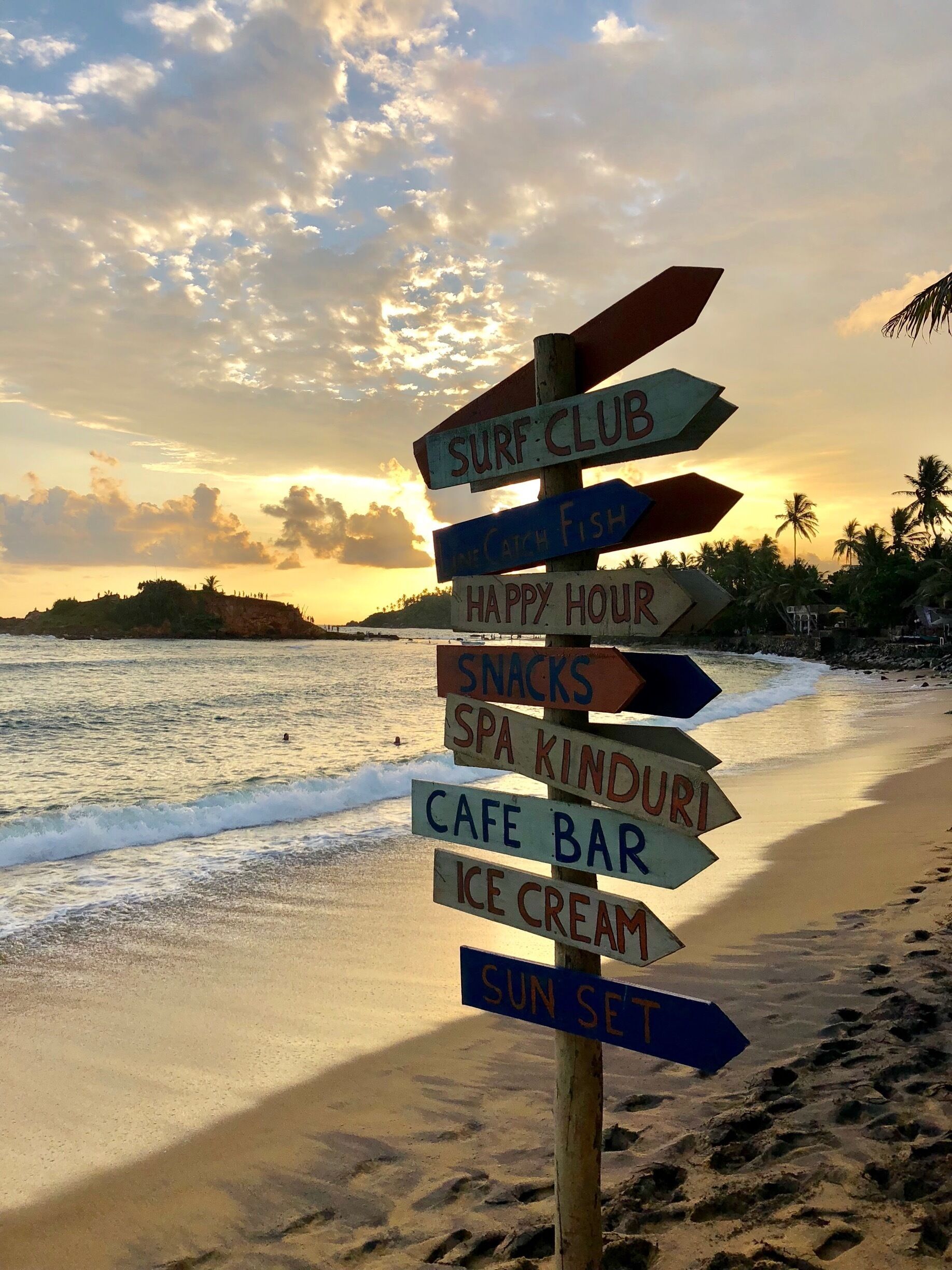 A beach with a signpost listing amenities like a surf club, happy hour, snacks, spa kinduri, cafe bar, ice cream, and sunset.