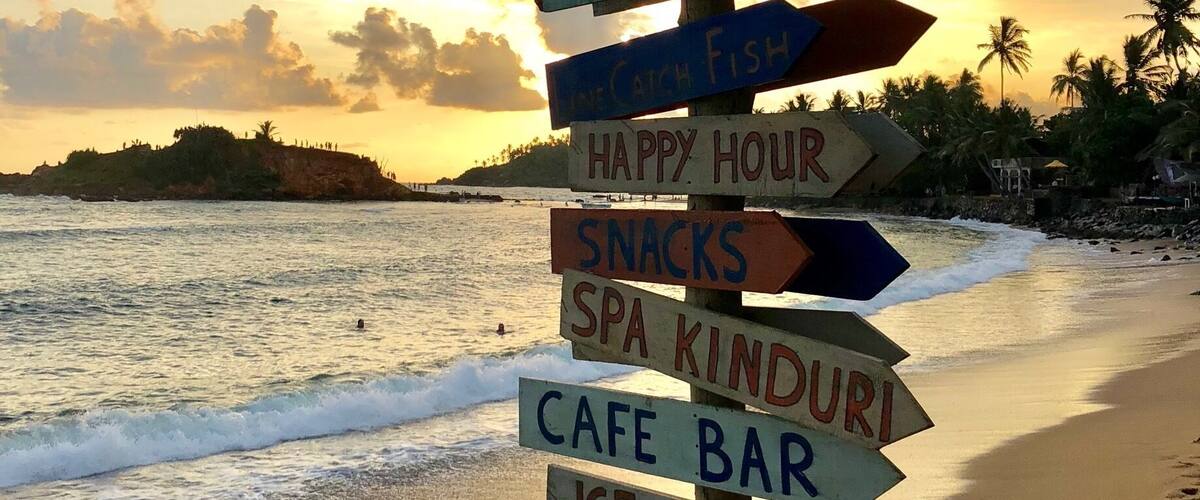 A beach with a signpost listing amenities like a surf club, happy hour, snacks, spa kinduri, cafe bar, ice cream, and sunset.