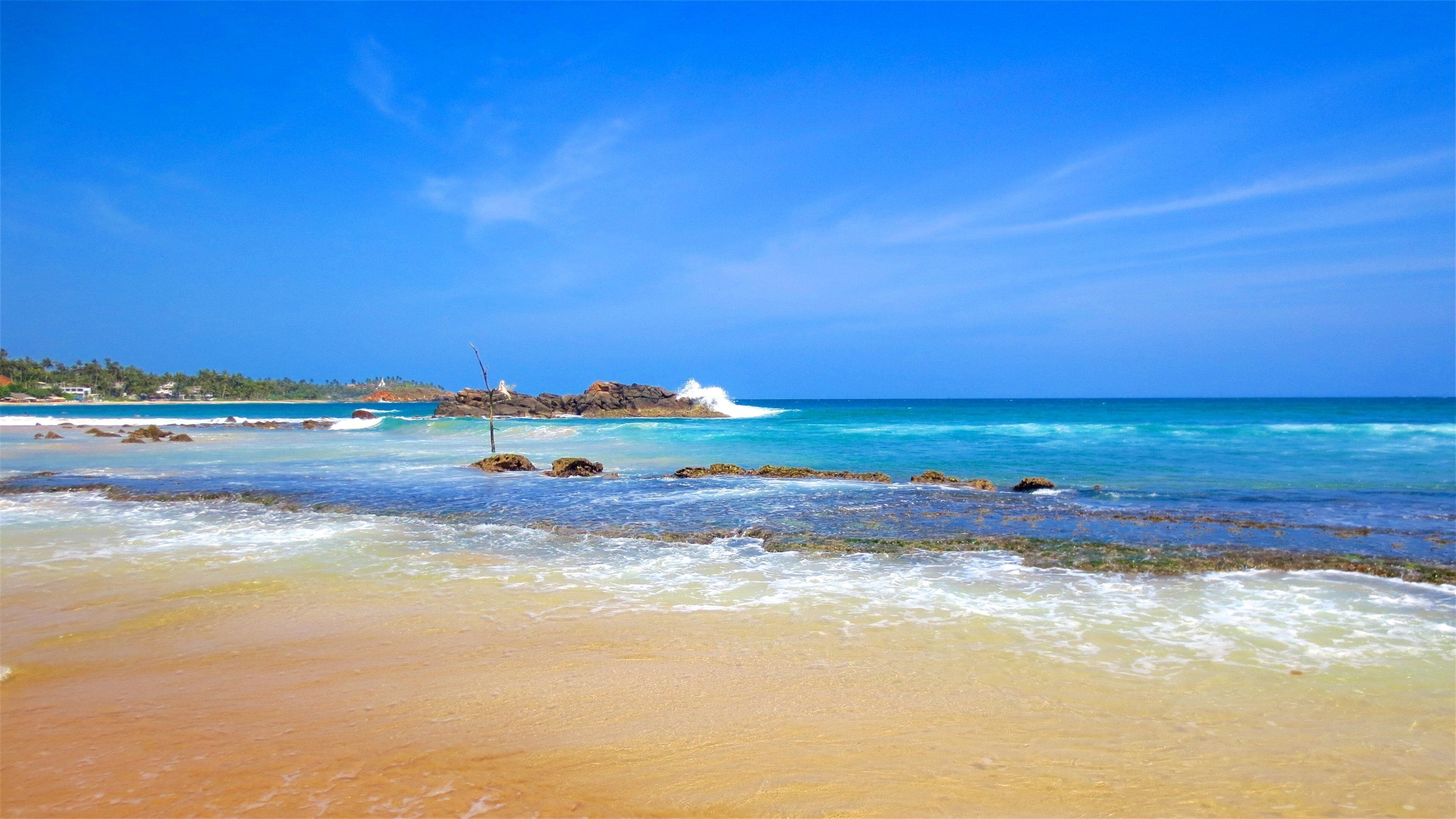 A beach with clear turquoise waters, rocky outcrops, and a distant shoreline with vegetation.