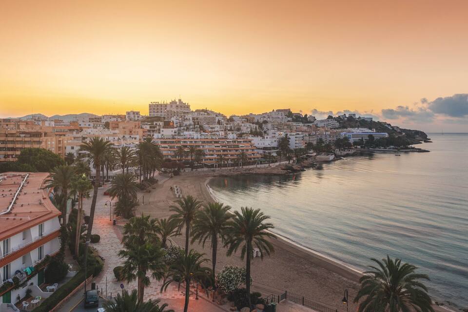 A coastal cityscape at sunset with buildings, a beach, and palm trees.