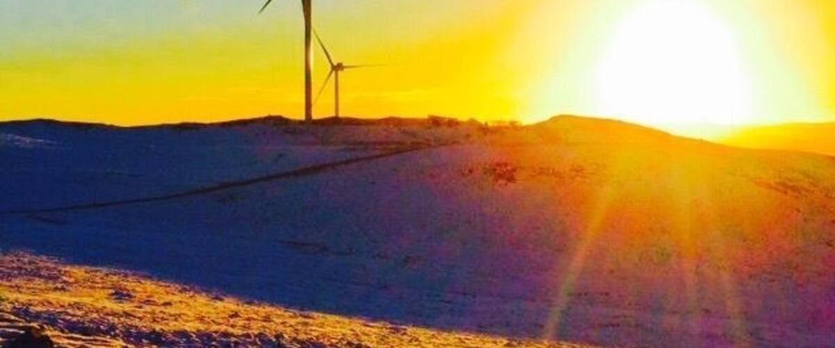 A wind turbine in a snowy landscape with the sun setting in the background.