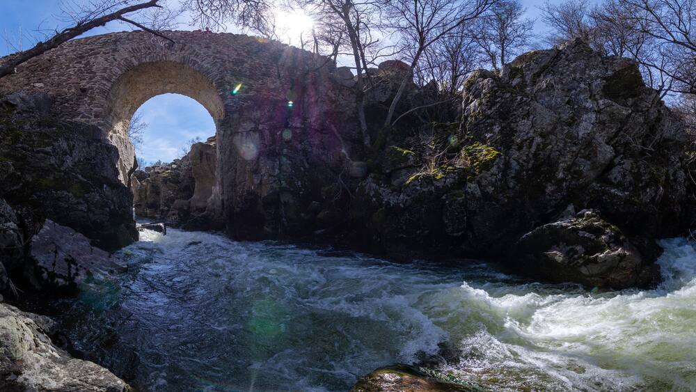 Un pont en pierre, dont l’arche s’étend au-dessus d’une rivière à courant rapide, est entouré d’un paysage rocheux et de quelques arbres clairsemés.