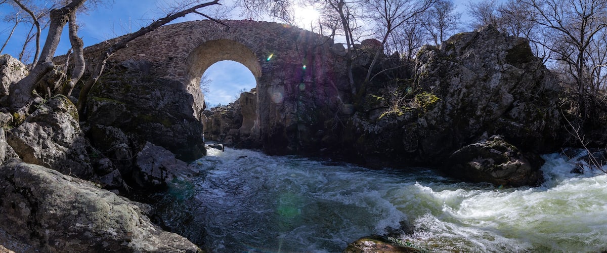A stone bridge arching over a fast-flowing river, surrounded by rocky terrain and sparse trees.