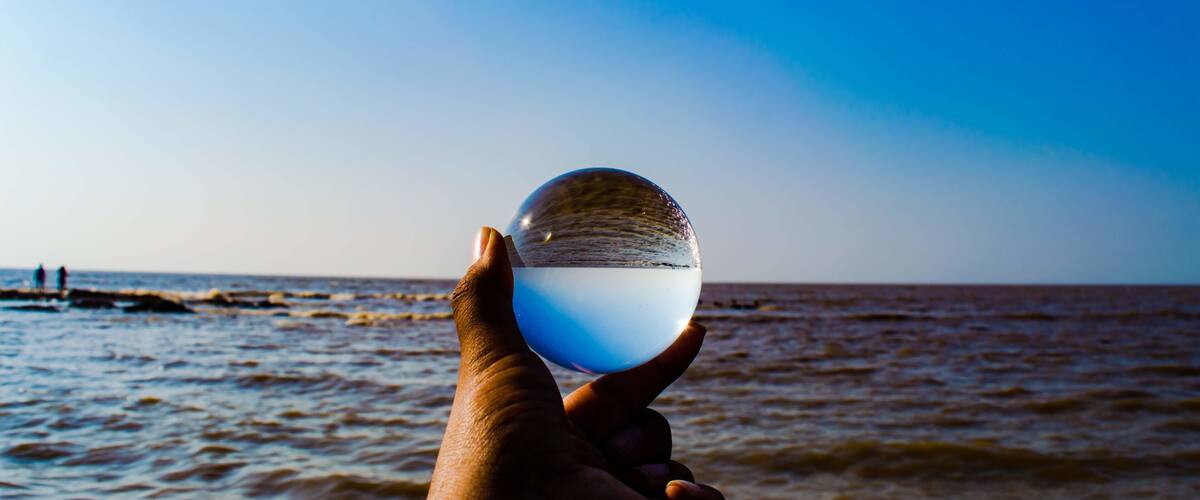A hand holding a clear crystal ball over a beach with waves and a clear sky.