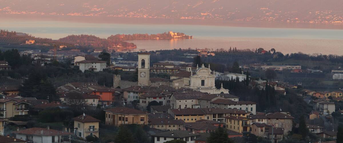 A town with historic buildings and a prominent church with a clock tower, overlooking a calm lake and a mountain range.