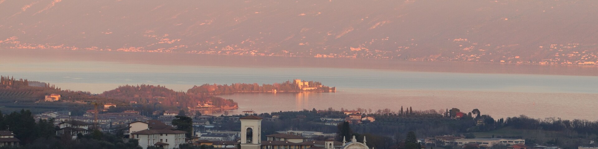 A town with historic buildings and a prominent church with a clock tower, overlooking a calm lake and a mountain range.