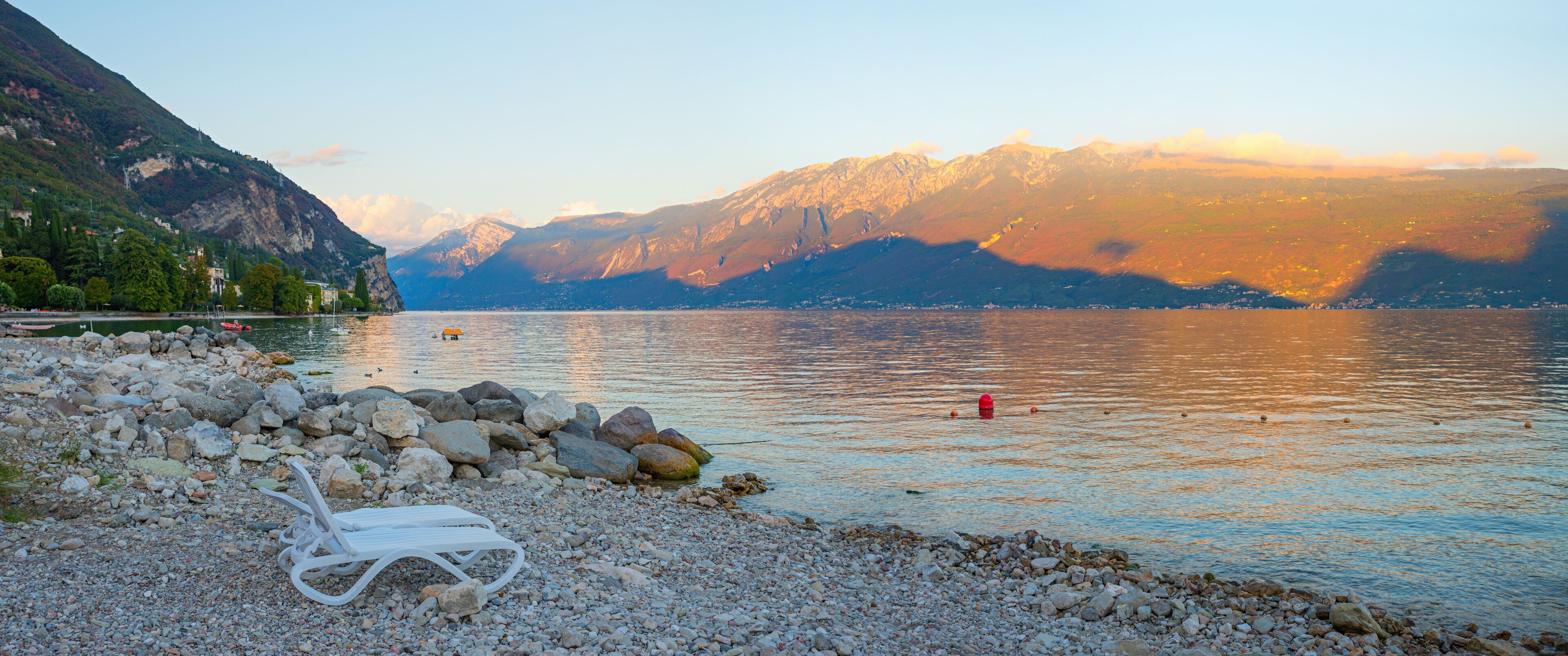 A lakeside view with a white lounge chair, rocky shore, and mountains in the background.