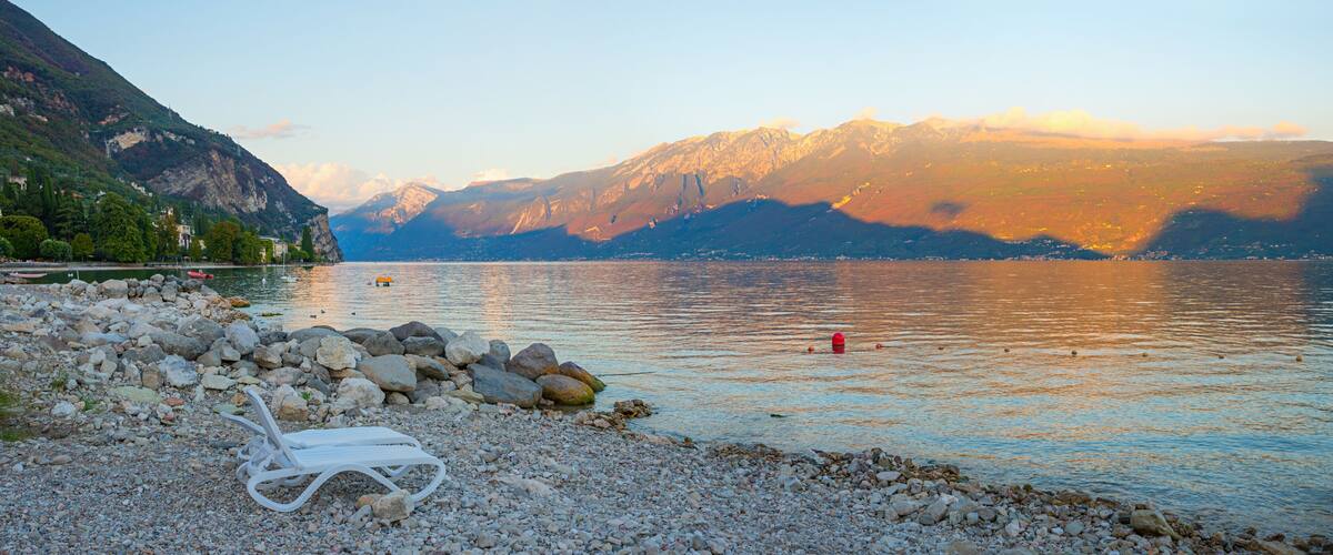 A lakeside view with a white lounge chair, rocky shore, and mountains in the background.