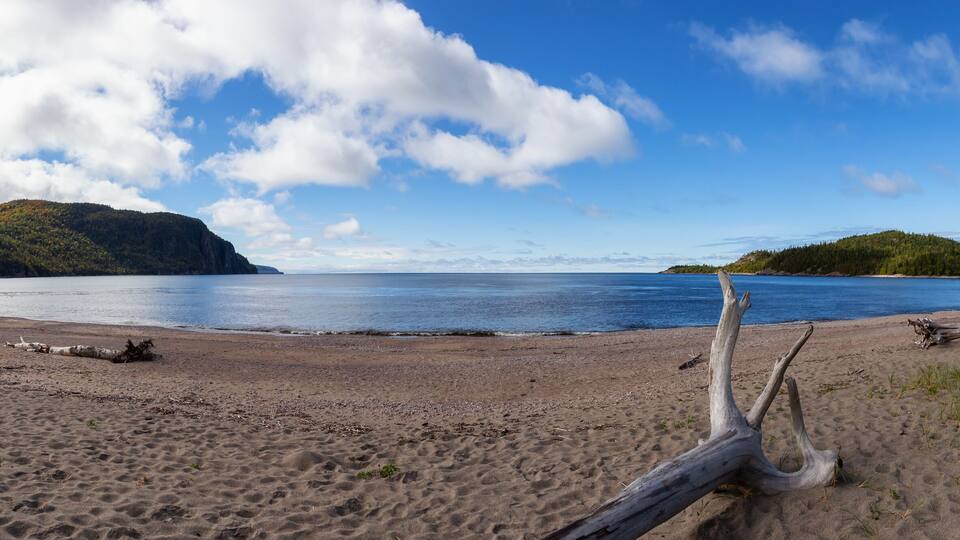 A sandy beach with driftwood and a calm lake surrounded by hills.