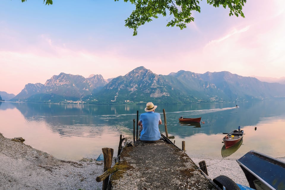 A man in a blue shirt and straw hat sitting on a dock overlooking a calm lake with mountains in the background.