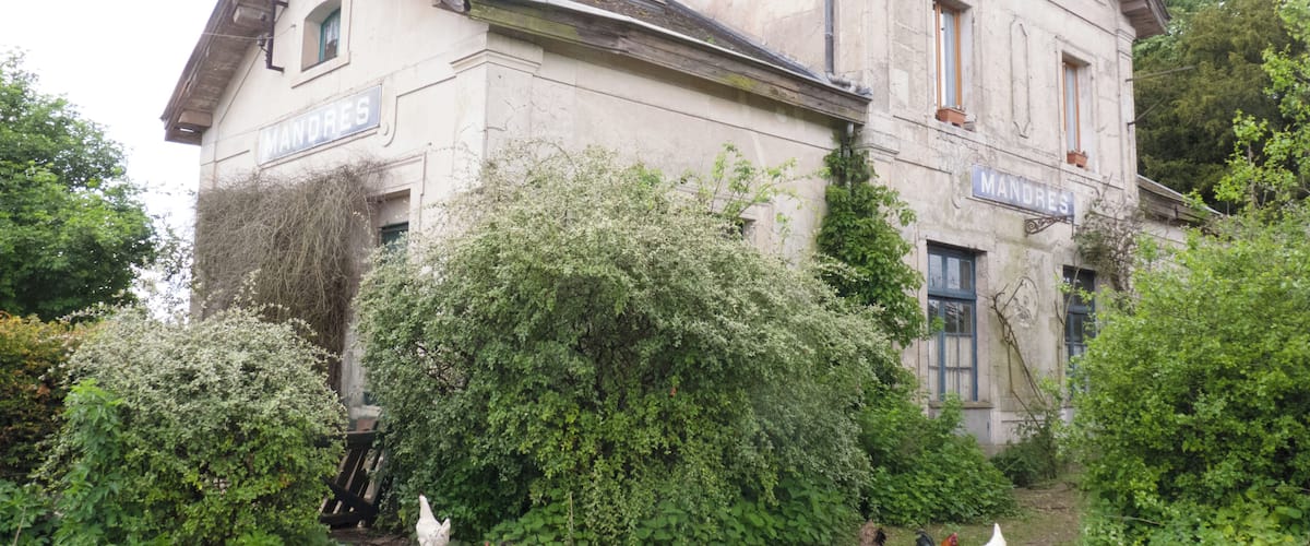 A historic building with the name 'MANDRES' on its facade, surrounded by lush greenery and chickens.
