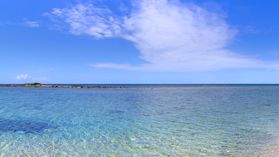 A clear blue sea with a sandy beach and a rocky outcrop in the distance.