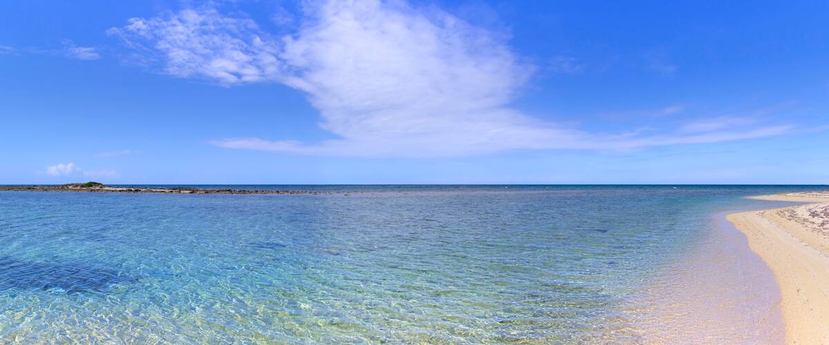 A clear blue sea with a sandy beach and a rocky outcrop in the distance.