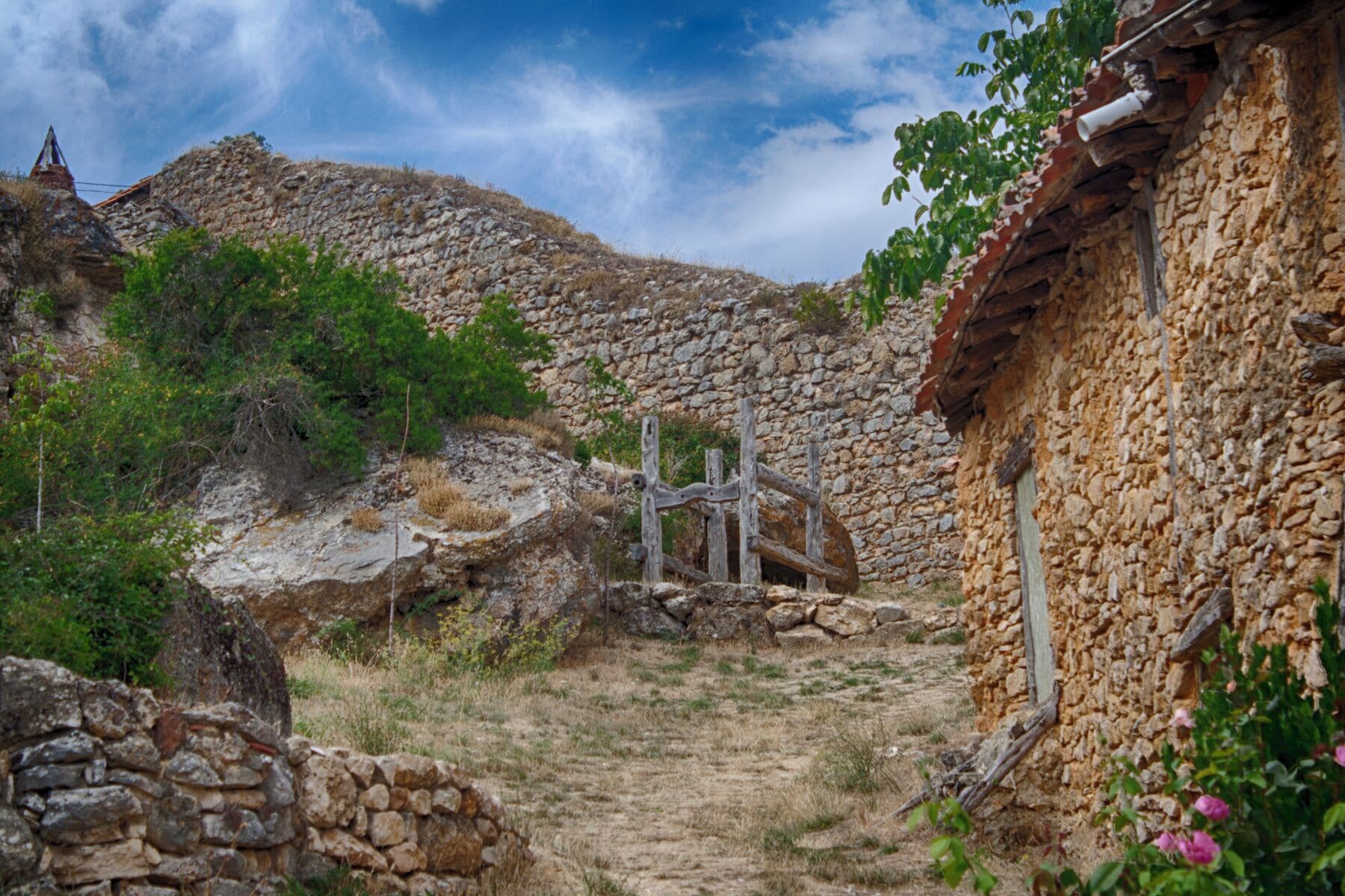 A rural stone pathway leading to a stone building with a tiled roof, surrounded by vegetation and a stone wall.
