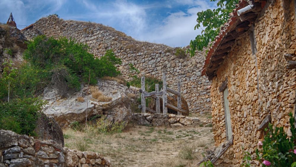 A rural stone pathway leading to a stone building with a tiled roof, surrounded by vegetation and a stone wall.