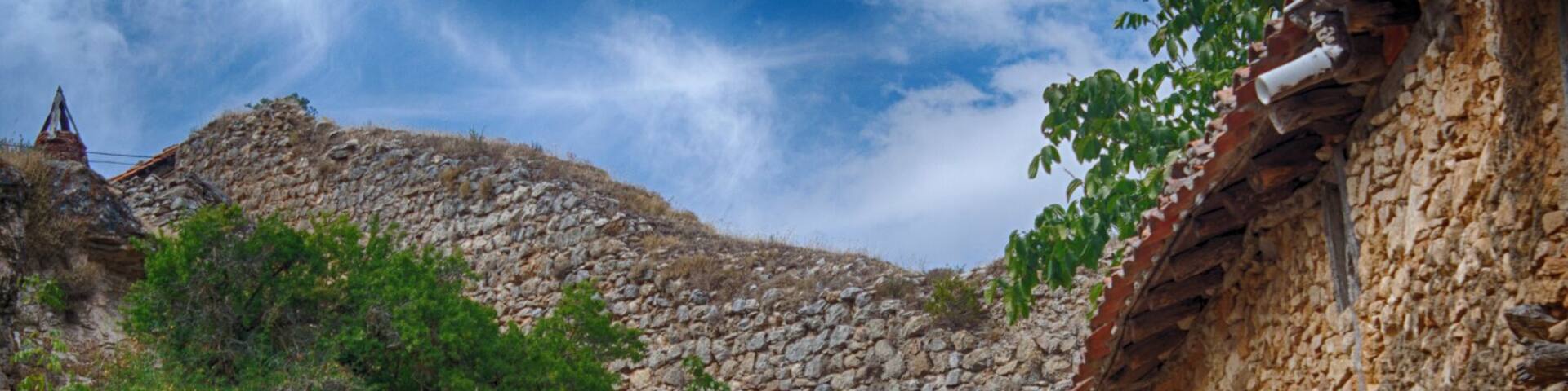 A rural stone pathway leading to a stone building with a tiled roof, surrounded by vegetation and a stone wall.