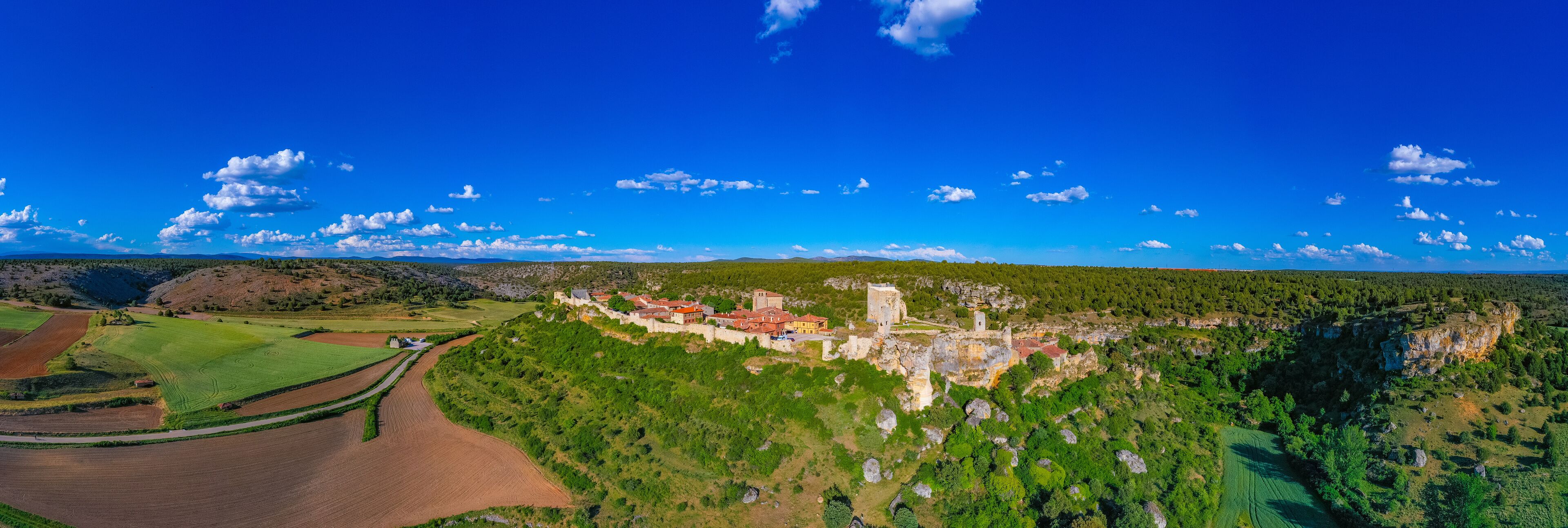 A panoramic view of a historic castle on a hill, surrounded by lush green fields and a clear blue sky.