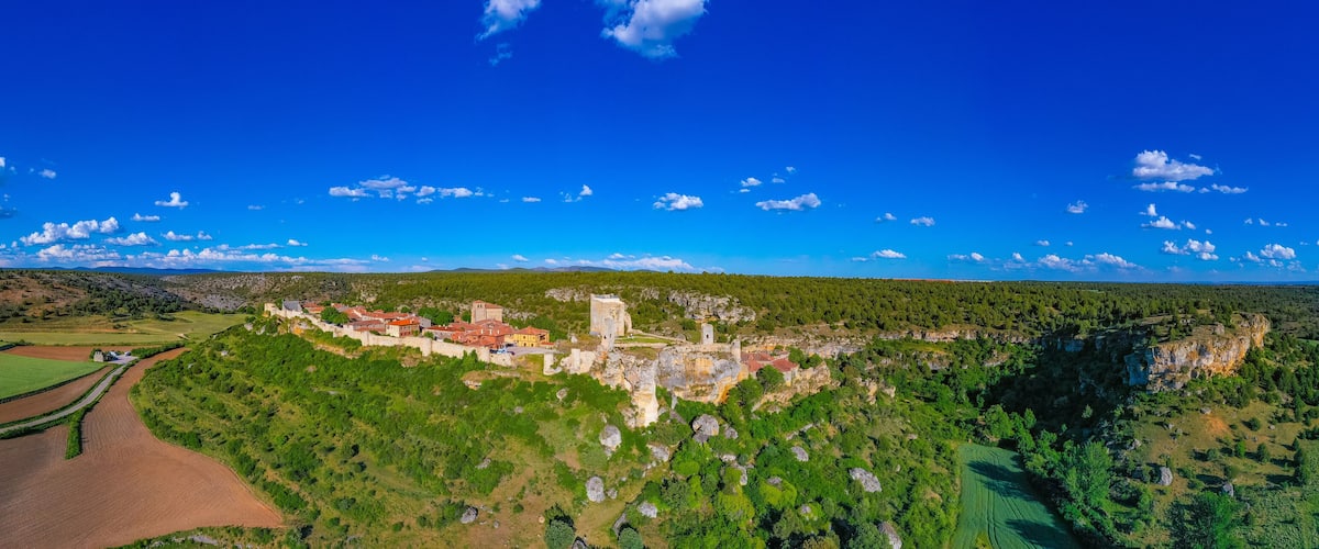 A panoramic view of a historic castle on a hill, surrounded by lush green fields and a clear blue sky.