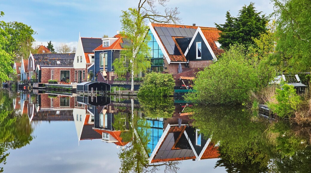 A picturesque scene showing a row of charming houses nestled next to a tranquil body of water, reflecting the clear blue sky and lush greenery