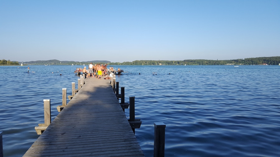 Eine Holzstege, der sich in einen stillen See erstreckt, mit Personen darauf und Booten in der Ferne.