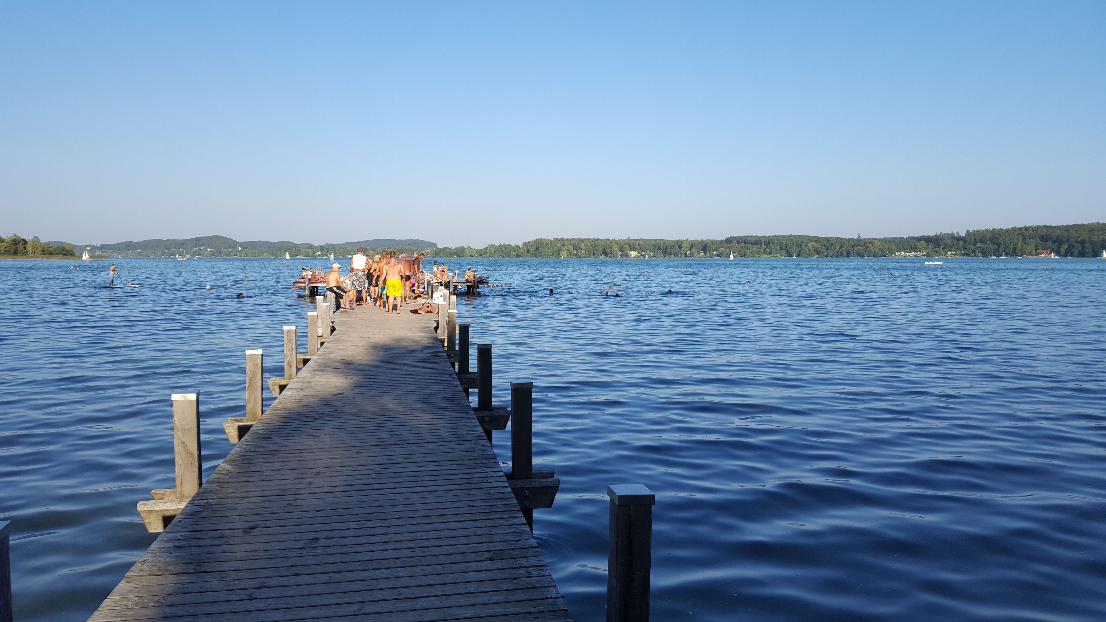 A wooden pier extending into a calm lake with people on it and boats in the distance.