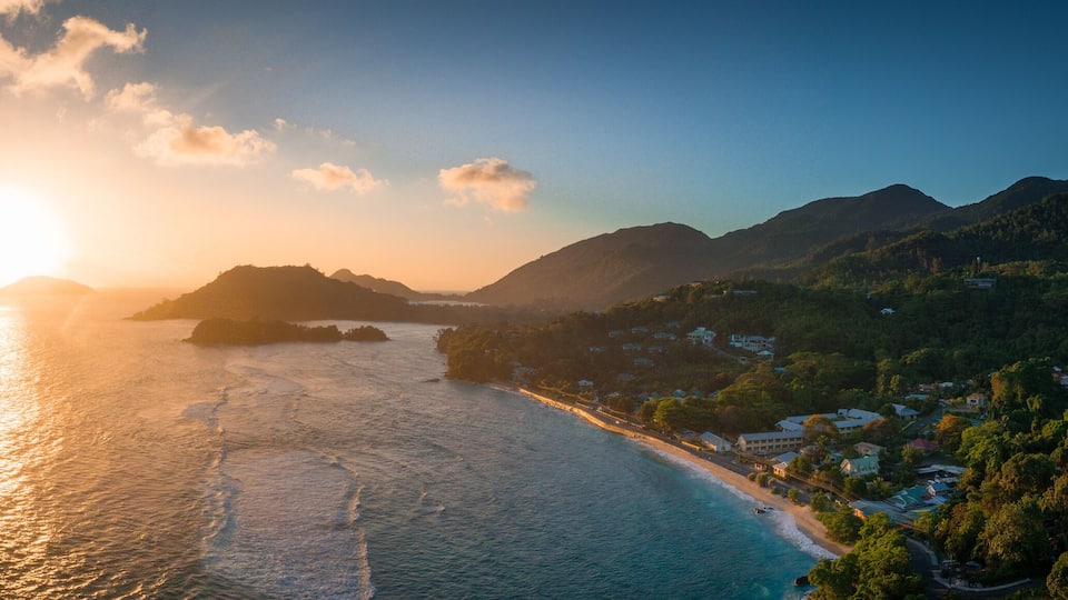 A coastal landscape with a beach, a sun setting over the sea, and a mountainous area with vegetation.
