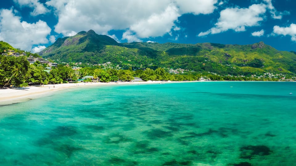A tropical beach with clear turquoise waters, a sandy shore, and lush green mountains in the background.