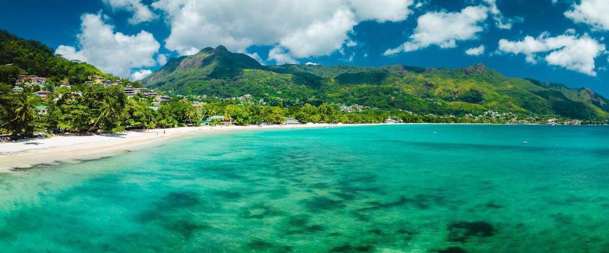 A tropical beach with clear turquoise waters, a sandy shore, and lush green mountains in the background.
