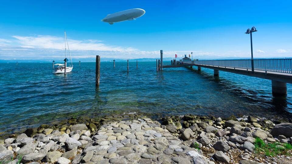 A pier extending into a calm body of water, a sailboat, and a blimp in the sky.