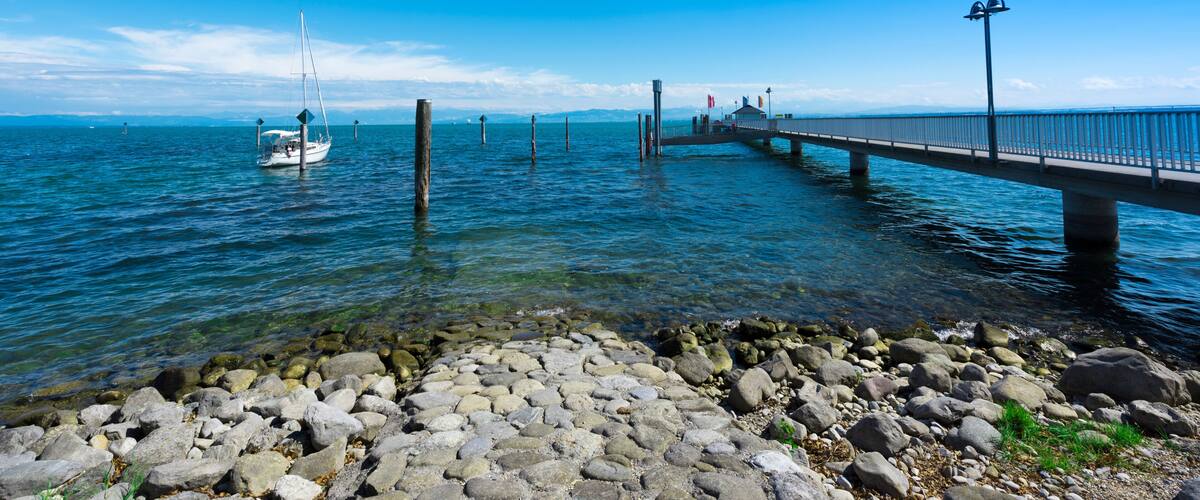 A pier extending into a calm body of water, a sailboat, and a blimp in the sky.