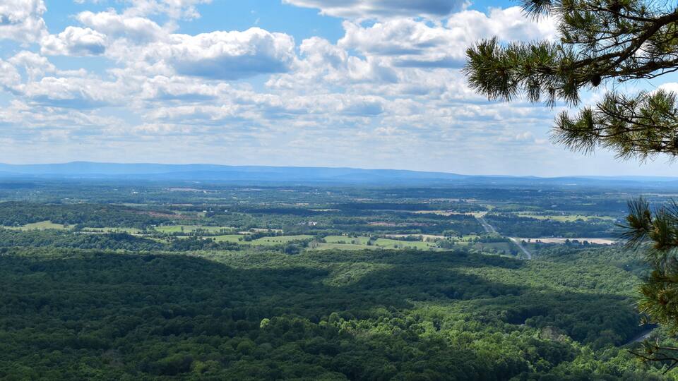 A panoramic view of a lush green landscape with a winding road, dense forests, and distant mountains under a partly cloudy sky.
