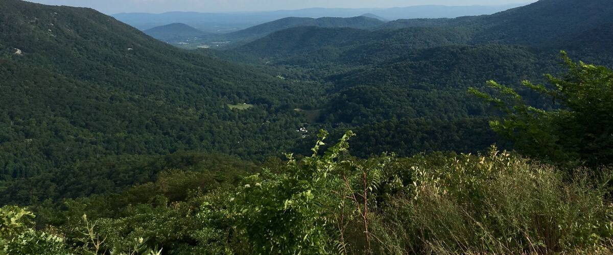 A mountainous landscape with dense forests and rolling hills under a partly cloudy sky.
