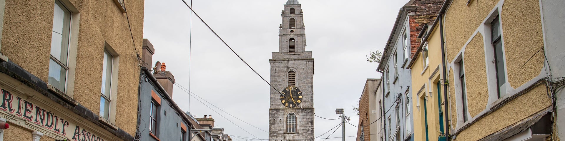A narrow street with historic buildings, a clock tower, and parked cars.
