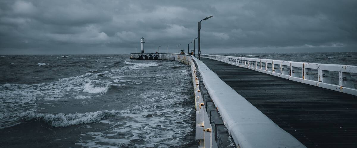 A pier extending into a stormy sea with a lighthouse in the distance.