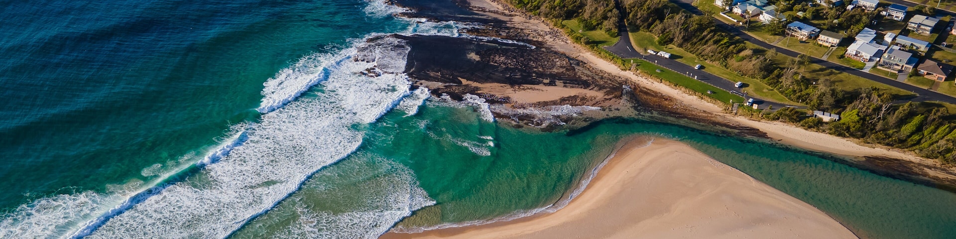 Vue aérienne d’une zone côtière avec une plage, des eaux d’un bleu limpide et un quartier résidentiel.