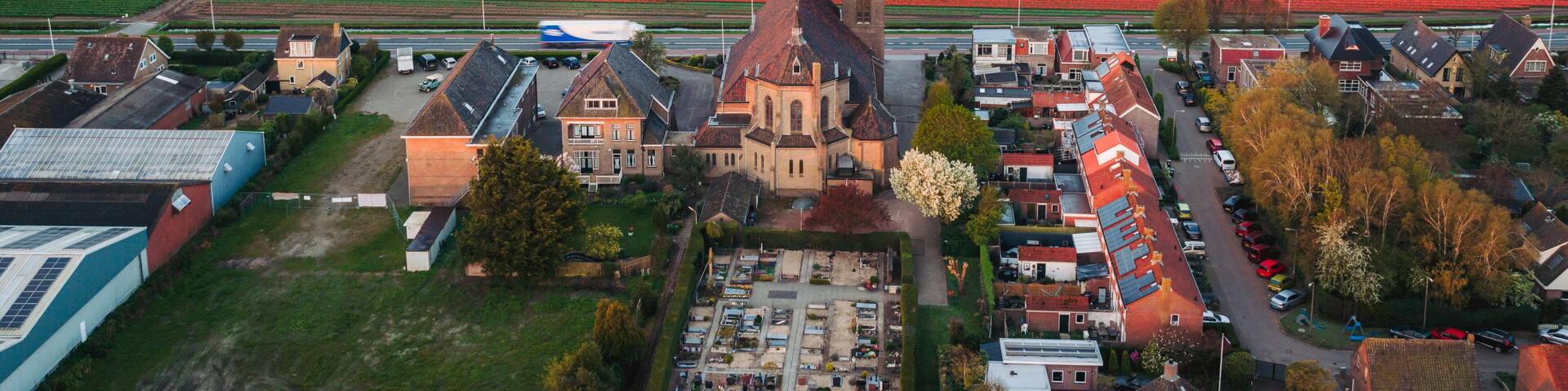 Aerial view of a historic church and cemetery surrounded by vibrant blooming tulip fields and village houses in De Zilk, Zuid-Holland, Netherlands.