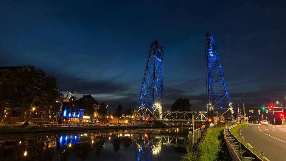 A nighttime view of a bridge with blue lighting, a calm river reflecting the lights, and a road with traffic lights.
